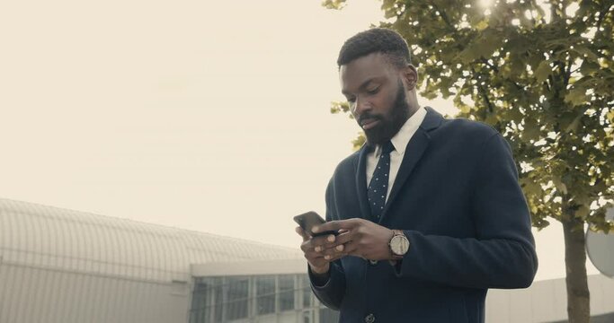 Attractive Afro American Businessman walking in the Street with Smartphone in his Hands. Working outdoor. Looking serious and Confident. Having nice Style and wearing luxury clothes.