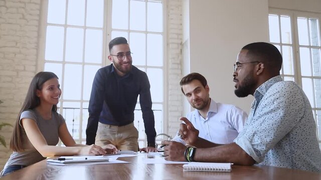 African male leader talking to caucasian coworkers group joking at group briefing, happy friendly diverse business team people listen to black coworker tell funny idea during corporate office meeting