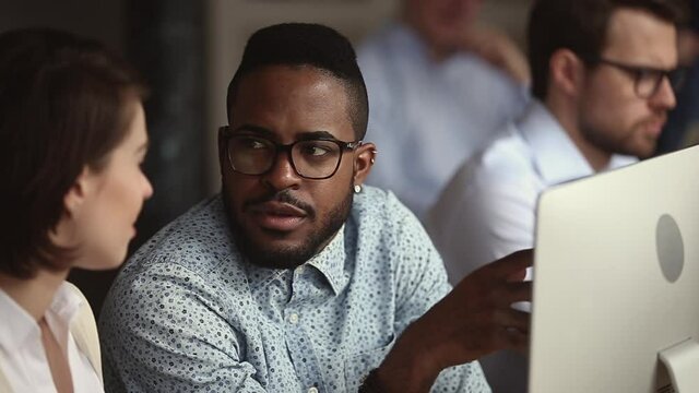 Male african black manager mentor teach help female caucasian intern trainee new employee explain work skills at workplace, serious diverse coworkers talk cooperate with computer in office apprentice