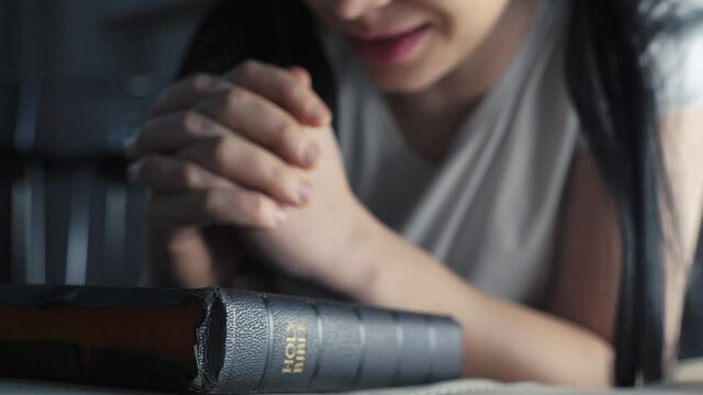 girl praying indoors at bedtime on bible. religion concept evening prayer woman lifestyle brunette hands on bible praying by bed