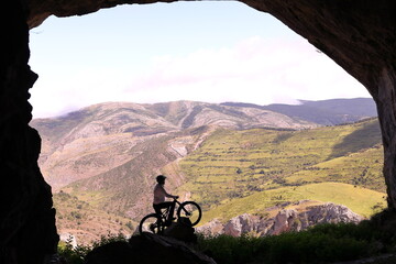 Fototapeta na wymiar BIKER WITH A MOUNTAIN BIKE AT THE ENTRANCE OF A LARGE CAVE AT THE TOP OF A LARGE VALLEY