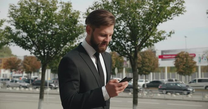 Video Shot of Handsome athletic Businessman Wearing formal Suit and having Smartphone. Slow motion of Attractive Man Typing on the Screen, Looking Happy and Pleasant. Business Man having a Walk.