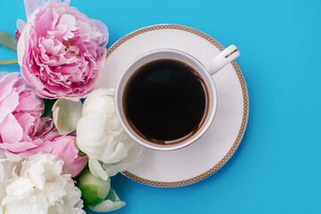 White cup of coffee and white and pink peony flowers on a blue background.Summer romantic breakfast.Good morning concept.Selective focus, top view