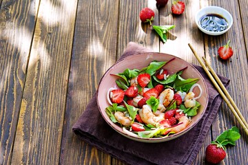 Salad with fresh strawberries, fried shrimp and young beet tops in a pink bowl on a wooden background.