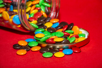 Frasco con confites de chocolate coloridos abierto y acostado sobre fondo infinito rojo 