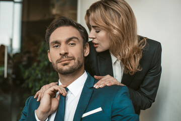 Passionate couple flirts at work. Office romance concept. Caucasian man and woman in love wearing formalwear. Man looks at camera, while woman whispers in his ear.