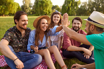 Fototapeta premium Group of friends sitting on grass, talking and high fiving in countryside. Joyful people enjoying time together outdoor in park