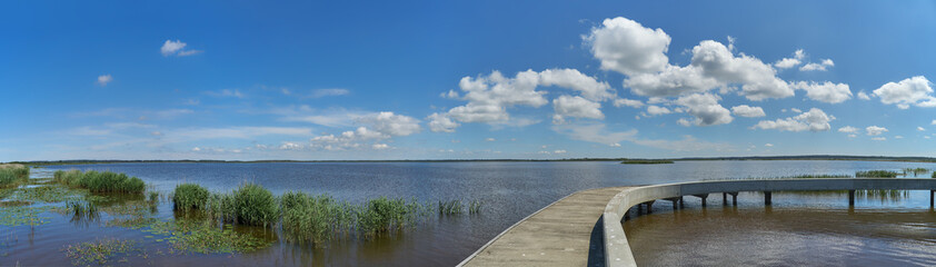 Scenic panorama of the lake Filsø in the "Naturpark Vesterhavet" (Denmark) on a sunny summer day with vivid blue sky and white clouds, in foreground are a round concrete jetty, reeds and water lilies