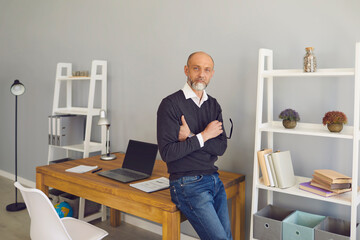 Portrait of serious mature man standing near his workplace with laptop indoors. Senior businessman in home office