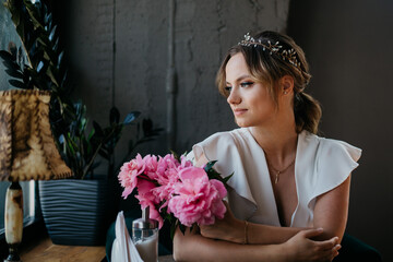 Young beautiful lady in white dress, with the bouquet of peonies, sitting in the cafe and looking to the window.