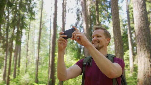 Tilt down panning shot of happy male adventure seeker with backpack walking among trees in forest and enjoying photographing nature on smartphone