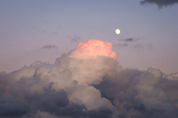 Top of a storm cloud turns pink after sunset. Bright moon shines in the air.