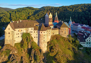 Fototapeta na wymiar Aerial view of Loket Castle, Burg Elbogen, gothic style castle on rocky cliff, surrounded by green hills. Massive fortification illuminated by setting sun. Karlovy Vary district, Czechia