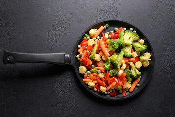 Frozen sliced colorful vegetables in frying pan on black background. View from above.