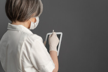 Caucasian woman in protective uniform using digital tablet. Gray-haired doctor works with electonic notepad. Close up shot. Rear or side view. Tinted image.