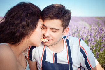 A young girl and a guy feed each other an orange in a lavender field. People in love sit against the background of purple lavender and eat. Love, love, happiness.