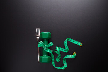 The fork is tied to a green dumbbell with a ruler against a dark background. The balance between nutrition and sport. Healthy lifestyle. Space for text. View from above.