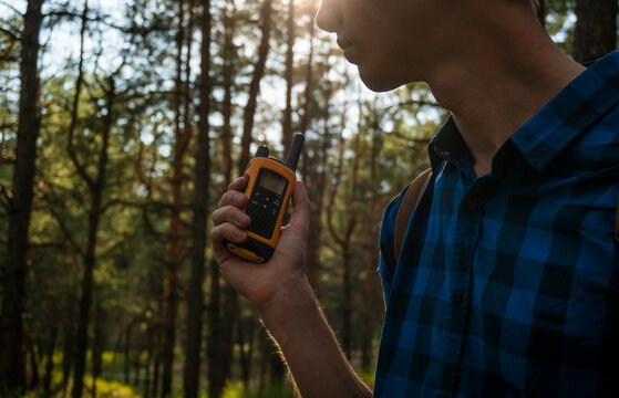 A young man in a blue plaid shirt with a walkie-talkie in his hand stands in a coniferous forest. - Powered by Adobe