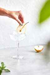 Classic martini with ice. Female hand decorates cocktail with pear slice. On a light background