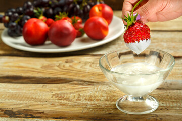 A female hand holds a strawberry in cream with a cup of whipped cream.