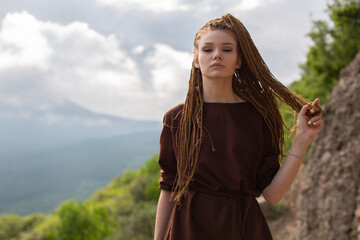 A red-haired girl with cornrows in a brown T-shirt-dress, defiantly looks into the camera holding a strand of hair in her hand and posing against the backdrop of a mountainside and wooded area