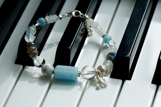 A handmade blue bracelet made of natural stones, aquamarine, cacholong, labrador, moonstone, with heart and key pendants, lies on black and white piano keys. Asymmetric arm bracelet