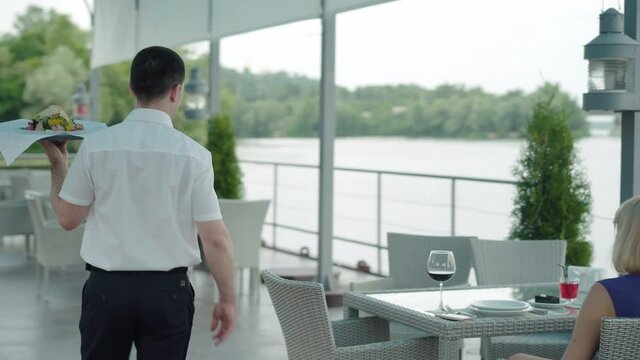 Camera follows professional male waiter bringing delicious dish to the table of two Caucasian women and leaving. Man serving food for clients in waterfront restaurant. Lifestyle, service, occupation.