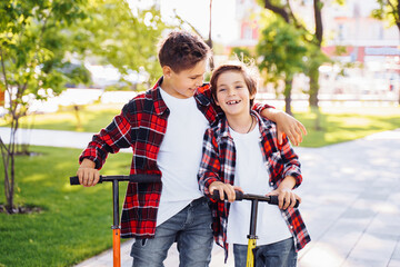 Two attractive european boys brothers, wearing red and white checkered flannel shirts, standing on scooters in the park. They laughing, smiling, hugging and having fun. Showing big fingers up sign