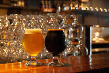 Two full beer glasses, on a counter at a bar, stack of glasses and beer taps in the background