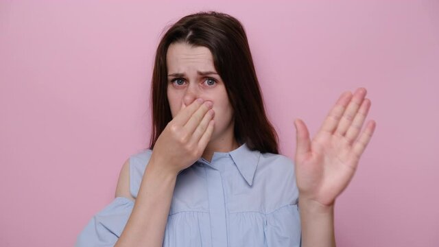 Confused displeased young caucasian woman covers nose with hand and showing stop gesture, dressed in blue shirt, isolated over pink wall background in studio. Repulsion to bad smell