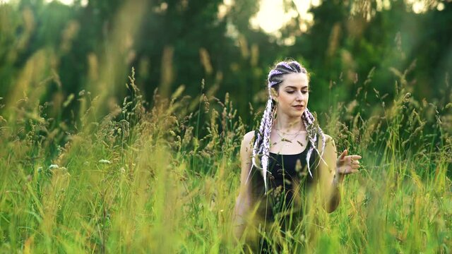 Young modern girl with colorful violet white blue braids. Braids, a black T-shirt and jeans. Stands in a field of green tall grass. Enjoys the nature. Depth of field. 4K resolution