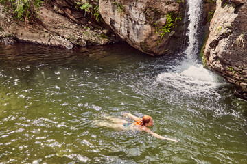 Young readhead woman swimming in mountain river in summer time with waterfall as background, Cerro Uritorco, Cordoba, Argentina, South America