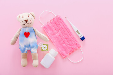 Protective medical mask with a bear toy, thermometer, and hand sanitizer on a pink background. Concept of quarantine of schools, home education