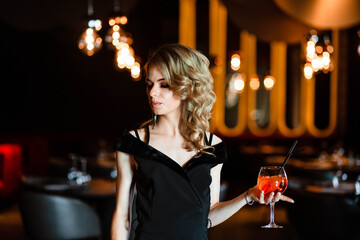 Beautiful blonde, young girl holding an aperol spritz. Cocktail aperol spritz in a glass. Portrait of a young girl in a restaurant. Portrait of a girl. Young girl in a restaurant with a glass.