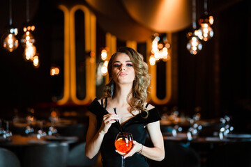 Beautiful blonde, young girl holding an aperol spritz. Cocktail aperol spritz in a glass. Portrait of a young girl in a restaurant. Portrait of a girl. Young girl in a restaurant with a glass.