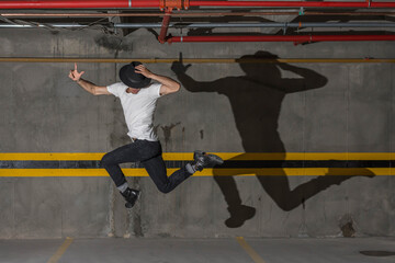 Young latin man with white t-shirt, hat and long shadow jumping in underground car parking