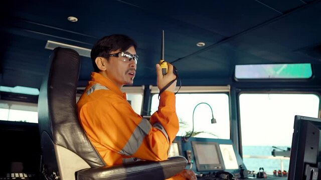 Filipino deck Officer on bridge of vessel or ship wearing coverall during navigaton watch at sea . He is speaking on GMDSS VHF radio, communication between vessels.