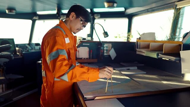 Filipino deck Officer on bridge of vessel or ship wearing coverall during navigaton watch at sea . He is plotting position on chart