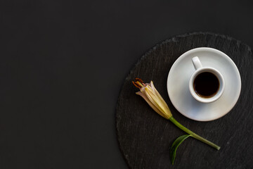 Cup of black coffee and fresh spring single bud of lily flower on a round slate on a black background. Romantic stylish elegance breakfast. Top view. Space for text.