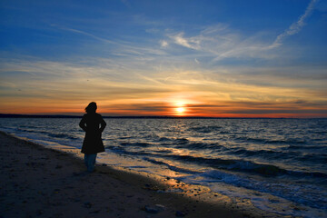 Fototapeta na wymiar A young woman watches the beautiful and colorful sunset at the West Meadow beach, Stony Brook, New York. 