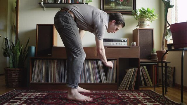 Barefooted hipster man walks up to a vintage stereo and puts on a vinyl record