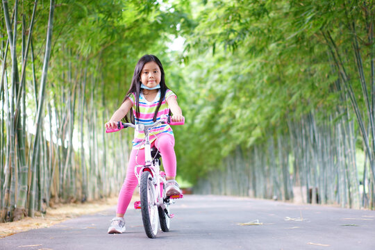 Asian Child Smile Or Kid Girl Wear Face Mask Under Chin And Cycling Fun On Bike Or People Bicycle On Public Park With Green Garden And Bamboo Tree For Sport Exercise To Healthy On Summer Relax Holiday