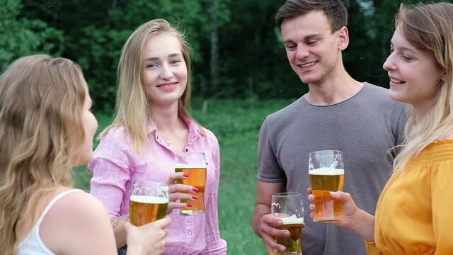Young beautiful Caucasian men girls relax on picnic in nature talk laugh, drink light beer from glasses. Friends at picnic in Park clink glasses with refreshing beverage. Active vacation in country