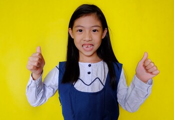 A cute young Asian girl in school uniform feeling happy, gesturing a 2 thumbs up sign in agreement. Bright yellow background.