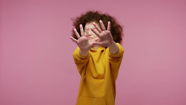 I'm afraid! Frustrated scared girl afro hairstyle in hoodie stretching out hands, stop gesture to hide from fear, looking terrified afraid, panic attack. indoor studio shot isolated on pink background