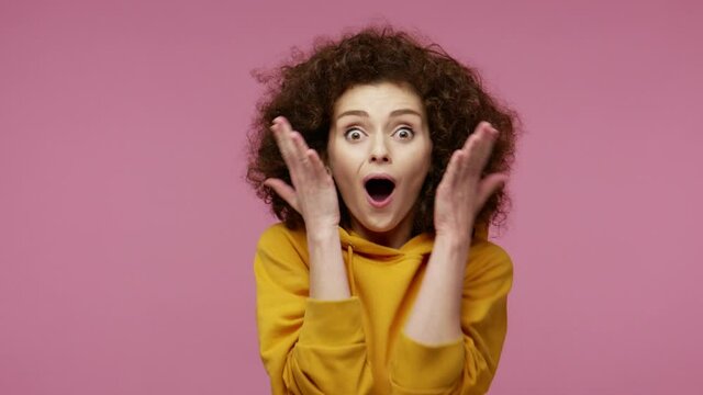Oh my god, wow! Amazed excited girl afro hairstyle in hoodie raising hands in surprise, looking at camera with big eyes, shocked by sudden victory. indoor studio shot isolated on pink background