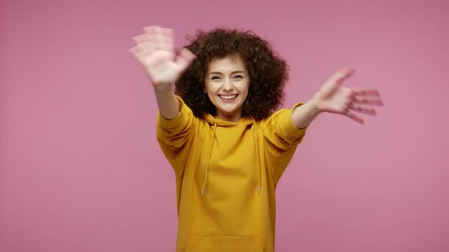 Sociable happy girl afro hairstyle in hoodie smiling friendly at camera and waving hands gesturing hello or goodbye, welcoming with hospitable expression. studio shot isolated on pink background