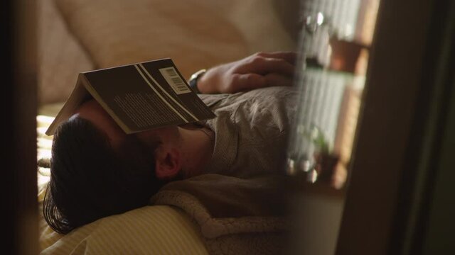 Man falls asleep and snores with a book over his head while laying on a bed