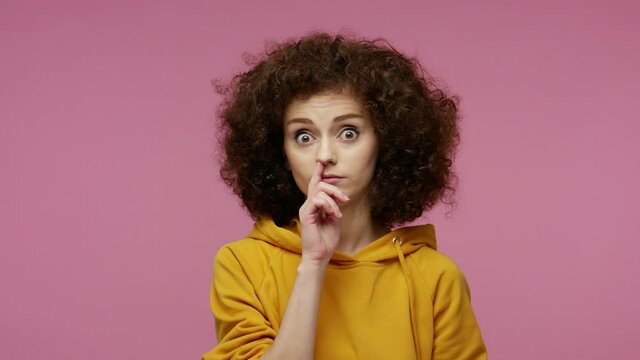 Weird funny girl afro hairstyle in hoodie picking nose, drilling boogers and looking around with comical stupid expression, bad manners, misconduct concept. studio shot isolated on pink background