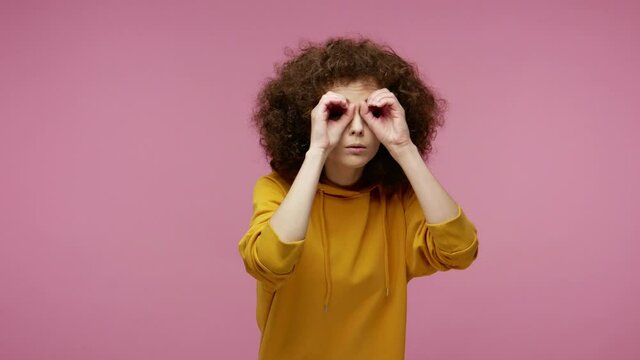 Curious girl afro hairstyle in hoodie looking through hands binocular gesture, zooming observing distance, expressing surprise shock happiness after seen discovery. indoor studio shot isolated